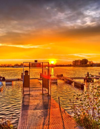 Sonnenuntergang über einem See mit Stegen, Booten und herbstlichen Büschen im Vordergrund