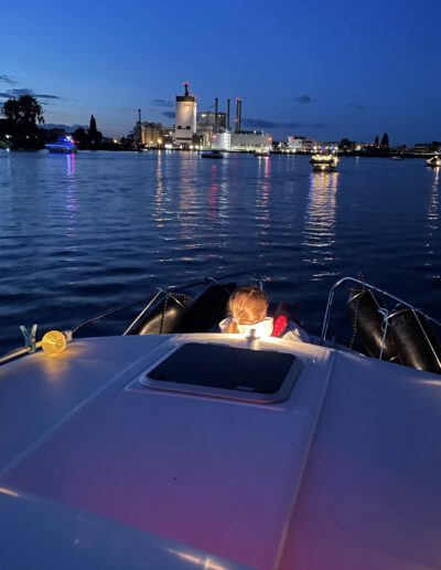 Person sitzt auf einem Boot bei Dämmerung, im Hintergrund beleuchtete Industrieanlagen am Flussufer