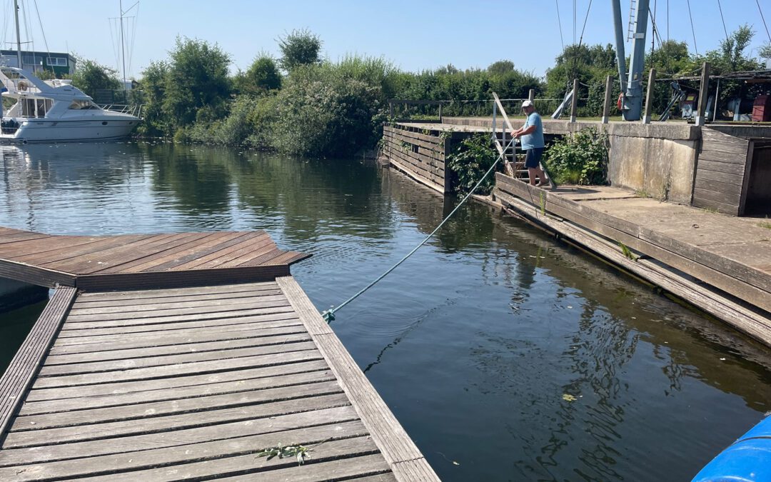 Holzsteg an einem ruhigen Wasserkanal mit Segelbooten, eine Person zieht ein Seil am Ufer