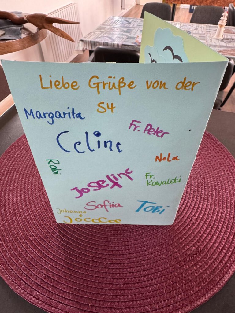 Klappkarte mit der Aufschrift 'Liebe Grüße von der S4' und mehreren handschriftlichen Namen in verschiedenen Farben auf einem runden, roten Tischset