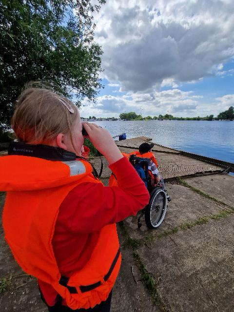 Zwei Kinder in orangen Schwimmwesten am Ufer eines Sees, einer sitzt im Rollstuhl, der andere schaut mit der Hand über den Augen in die Ferne
