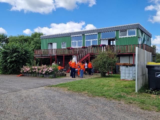 Grünes zweistöckiges Gebäude mit Balkon, davor Gruppe von Menschen in orangefarbener Kleidung, umgeben von Blumen und Büschen