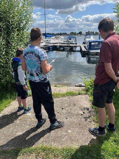 Drei Personen am Ufer eines Sees, zwei Jungen angeln mit einer Angelrute, ein Mann beobachtet sie, im Hintergrund Boote auf dem Wasser