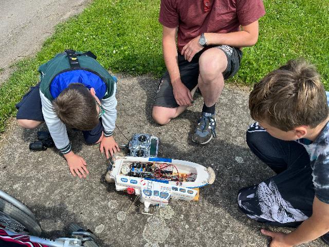 Drei Personen knien auf dem Boden und betrachten ein ferngesteuertes Modellboot mit sichtbarer Elektronik